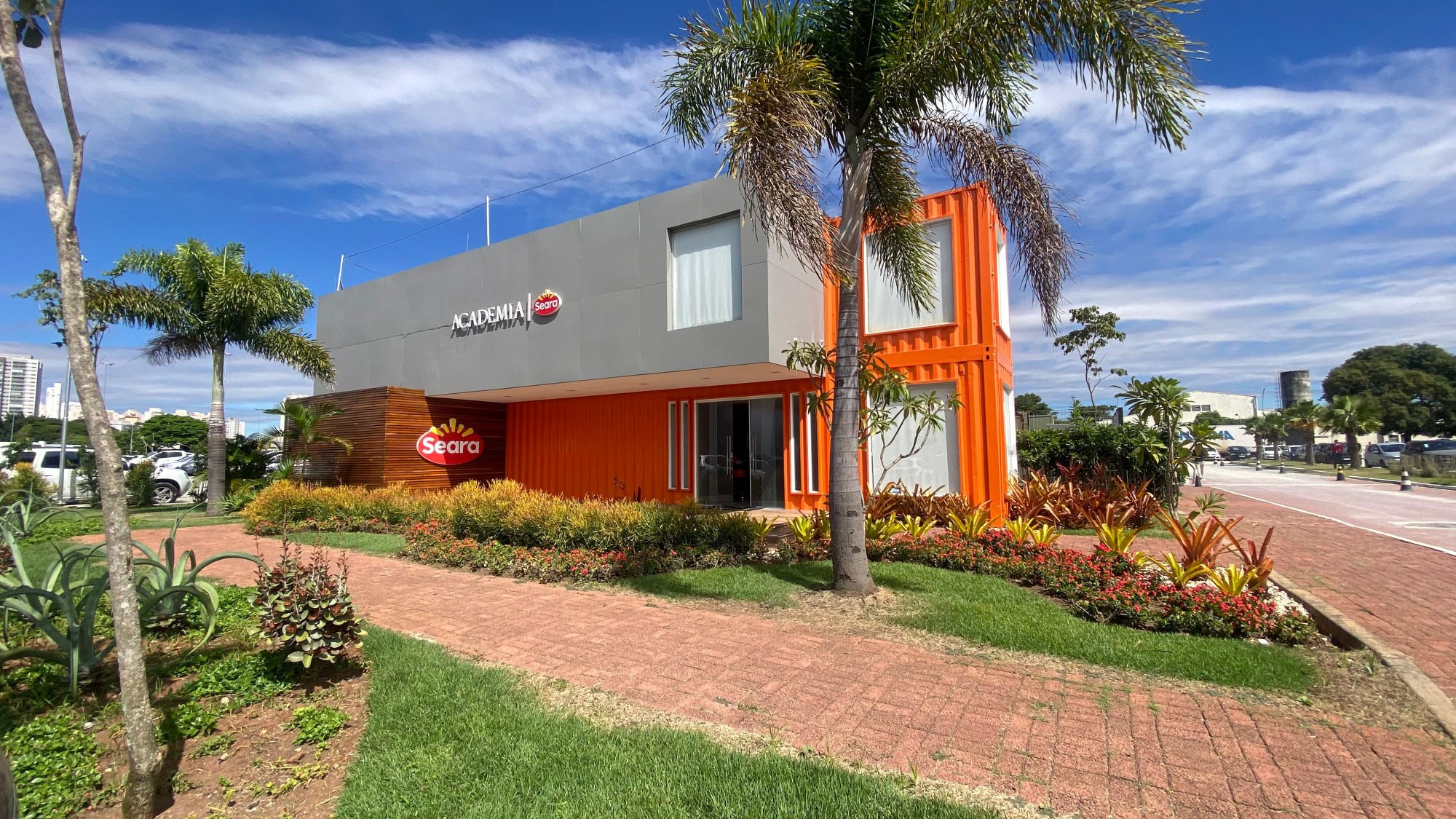 A modern two-story building in shades of gray and orange, with some wood accents, featuring the Seara brand logo, situated in a tree-lined parking lot on a sunny day.
